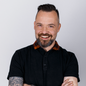 Porträt eines lächelnden Mannes mit Bart und Tattoos in einem schwarzen Hemd.